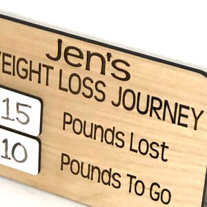 May include: A wooden sign with the text "Jen's Weight Loss Journey" and two white squares with the numbers "15" and "10" written on them. The text "Pounds Lost" and "Pounds To Go" are also written on the sign.