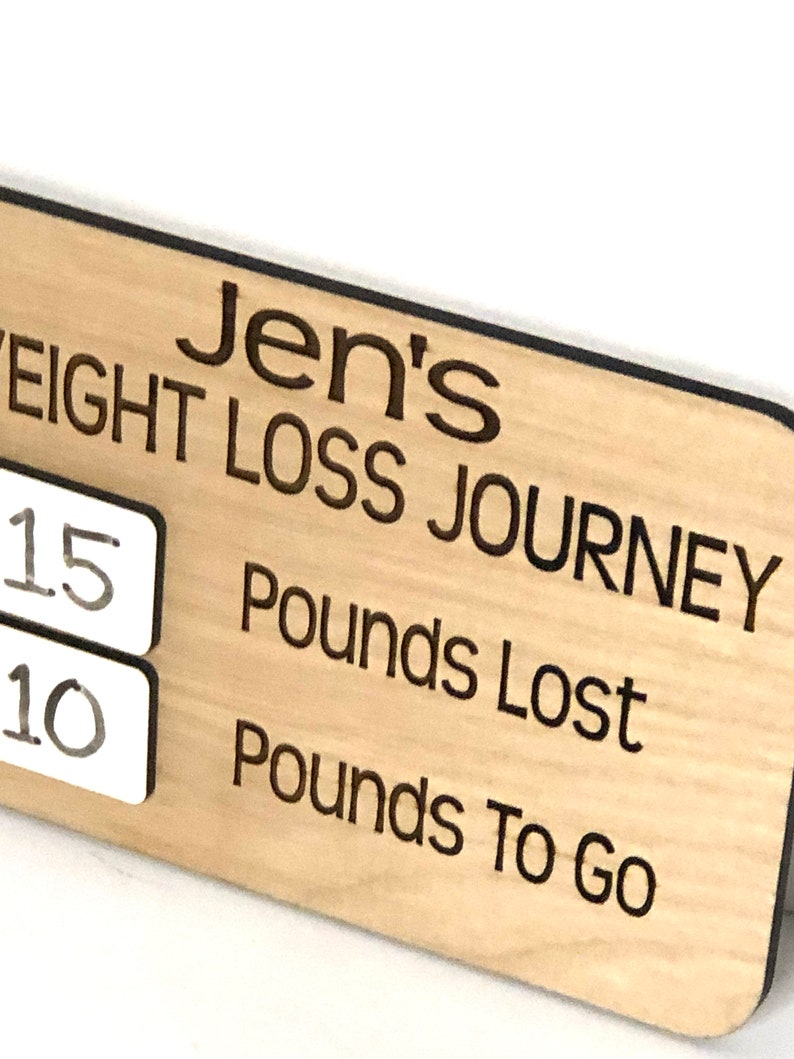 May include: A wooden sign with the text "Jen's Weight Loss Journey" and two white squares with the numbers "15" and "10" written on them. The text "Pounds Lost" and "Pounds To Go" are also written on the sign.