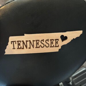 May include: A light brown wooden cutout of the state of Tennessee with the word "TENNESSEE" engraved in black lettering. A heart shape is cut out of the state outline.