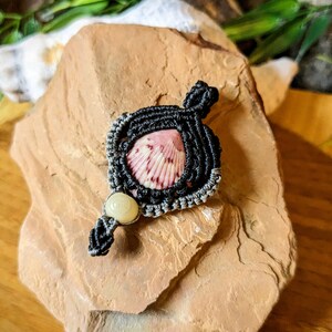 May include: A black and gray macrame knot pendant with a pink and white seashell in the center. A white bead hangs from the bottom of the pendant.