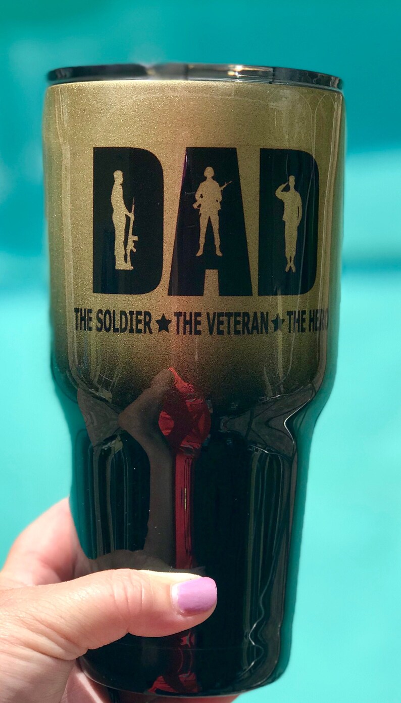 May include: A gold and black tumbler with the word "DAD" in large letters. The word is made up of three silhouette figures of soldiers. The text "THE SOLDIER THE VETERAN THE HERO" is printed below the word "DAD".