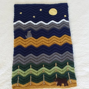 May include: A crocheted blanket with a night sky scene featuring a yellow moon, stars, and a wolf. The blanket also features a mountain range, trees, and a brown bear.