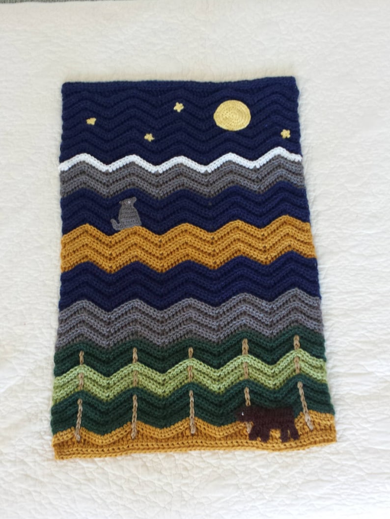 May include: A crocheted blanket with a night sky scene featuring a yellow moon, stars, and a wolf. The blanket also features a mountain range, trees, and a brown bear.