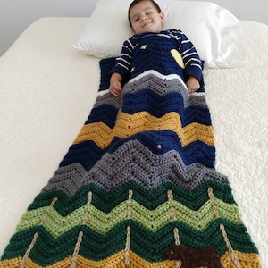 May include: A child is lying on a white bed with a blue, green, and yellow chevron patterned blanket. The blanket features a brown bear and trees.