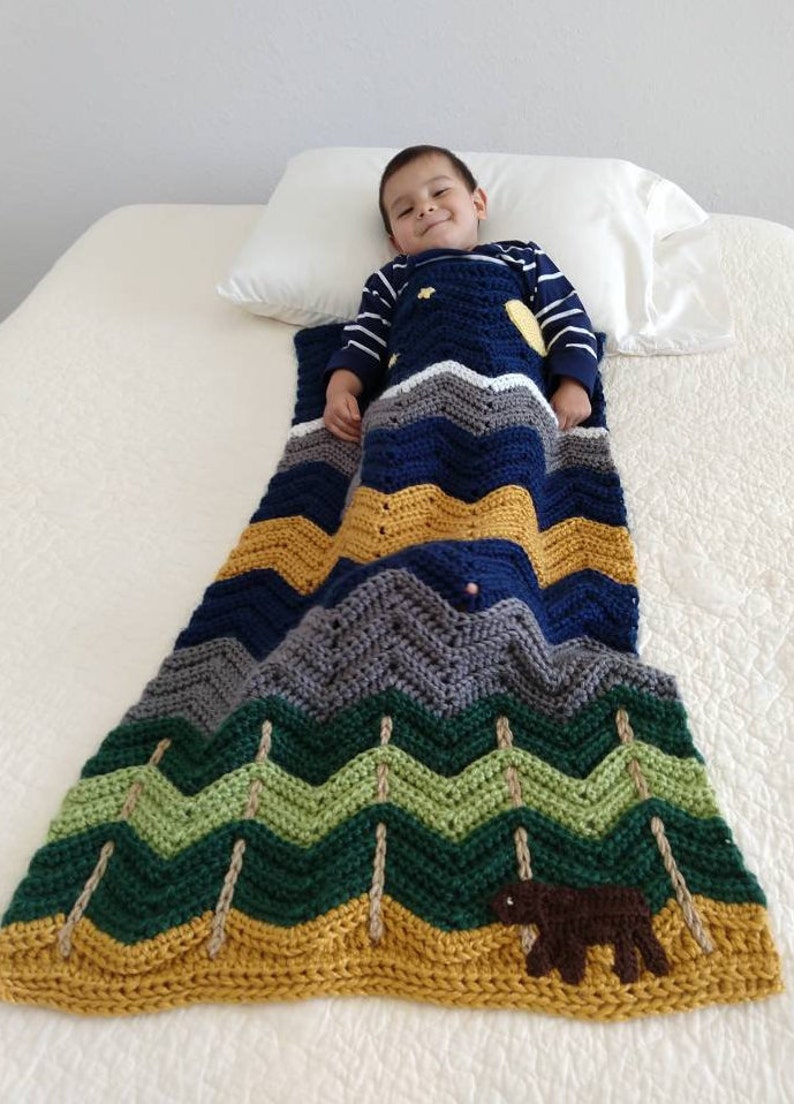 May include: A child is lying on a white bed with a blue, green, and yellow chevron patterned blanket. The blanket features a brown bear and trees.