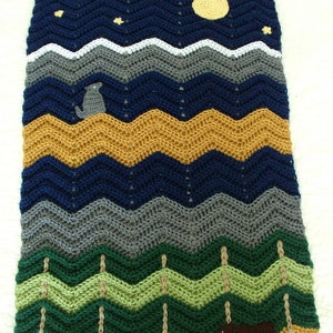 May include: A crocheted baby blanket with a woodland theme. The blanket features a blue night sky with stars and a yellow moon, a gray mountain range, green trees, and a brown bear and wolf.