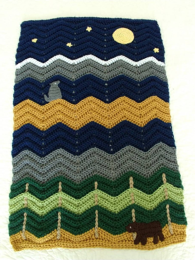 May include: A crocheted baby blanket with a woodland theme. The blanket features a blue night sky with stars and a yellow moon, a gray mountain range, green trees, and a brown bear and wolf.