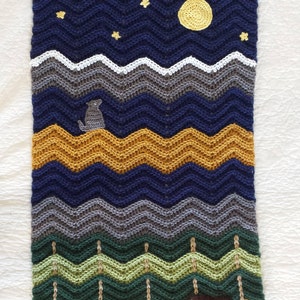 May include: Crocheted baby blanket with a blue, gray, green, and yellow chevron pattern. The blanket features a crocheted moon, stars, a bear, and a dog.