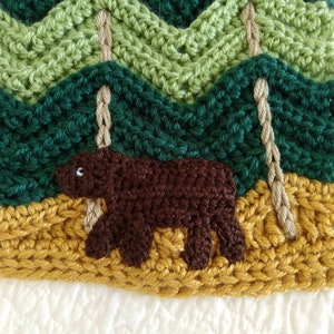 May include: Crocheted woodland scene with a brown bear in the foreground, green trees in the background, and a yellow crocheted ground.