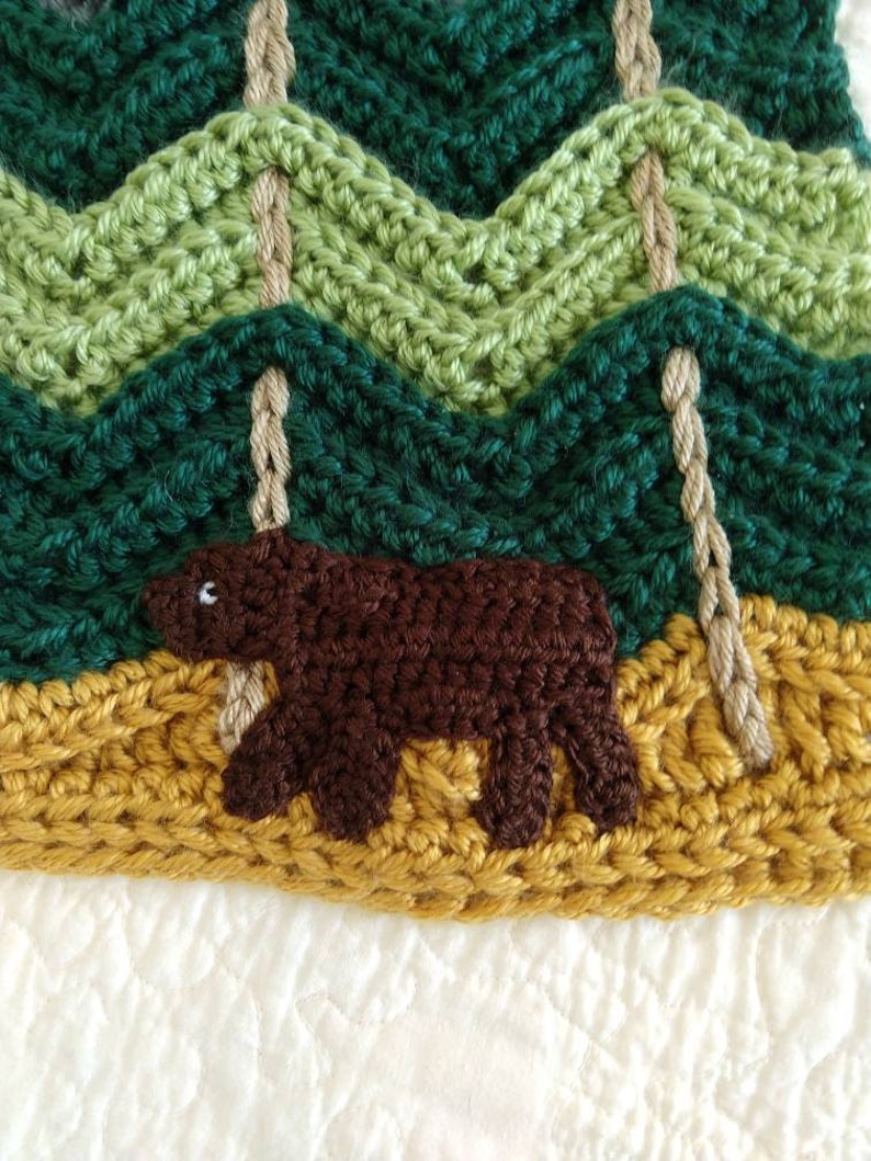 May include: Crocheted woodland scene with a brown bear in the foreground, green trees in the background, and a yellow crocheted ground.