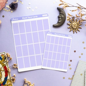 May include: A still-life arrangement featuring tarot cards, blank tarot card sheets, crystals, a candle, and celestial-themed decor on a lavender surface. The blank cards have a grid design. Dried flowers and gold sequins add detail.