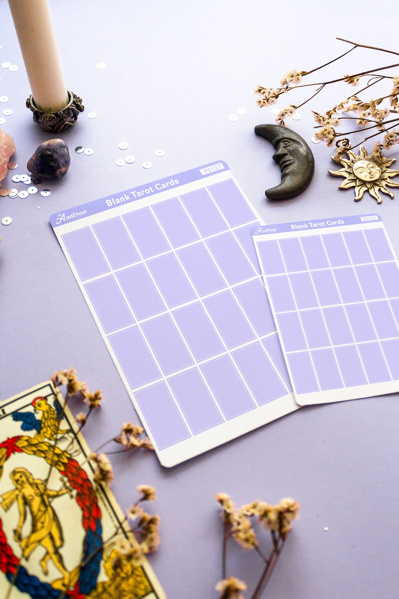 May include: Two lavender-colored blank tarot card sheets with grid layouts, labeled "Blank Tarot Cards" and "Aastrae". A vintage tarot card, a bronze moon, and a sun charm are also visible. The background is a light purple.