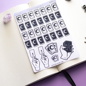 May include: A sheet of stickers featuring tarot card designs with a crescent moon and stars. The stickers are printed on white paper and have a black and white color scheme. The sheet also includes a hand holding a tarot card with a pentagram.