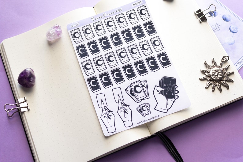 May include: A sheet of stickers featuring tarot card designs with a crescent moon and stars. The stickers are printed on white paper and have a black and white color scheme. The sheet also includes a hand holding a tarot card with a pentagram.