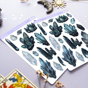 May include: Two sheets of stickers featuring watercolor illustrations of dark blue and clear crystals. The stickers are labeled "Dark Fairy Crystals" and include the brand name "Astron". Decorative elements include dried flowers, a crescent moon, and a sun charm.
