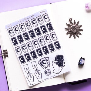 May include: A sheet of stickers featuring tarot card designs. The stickers are white with black outlines and feature a crescent moon, stars, and a hand holding a tarot card. The text "Tarot Cards #01" and "astrae.etsy.com" are printed on the sheet.