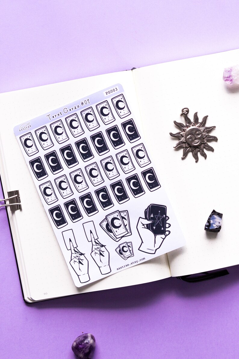 May include: A sheet of stickers featuring tarot card designs. The stickers are white with black outlines and feature a crescent moon, stars, and a hand holding a tarot card. The text "Tarot Cards #01" and "astrae.etsy.com" are printed on the sheet.