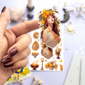 May include: A sticker sheet with a whimsical illustration of a woman with a crown of oranges and leaves. The sheet also includes smaller illustrations of oranges, candles, and other decorative elements.