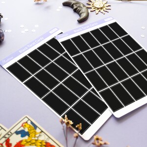 May include: Two sheets of blank tarot cards with black rectangles, labeled "Blank Tarot Cards" in white text. The cards are on a light purple surface, with decorative elements including a crescent moon, sun, and dried flowers.
