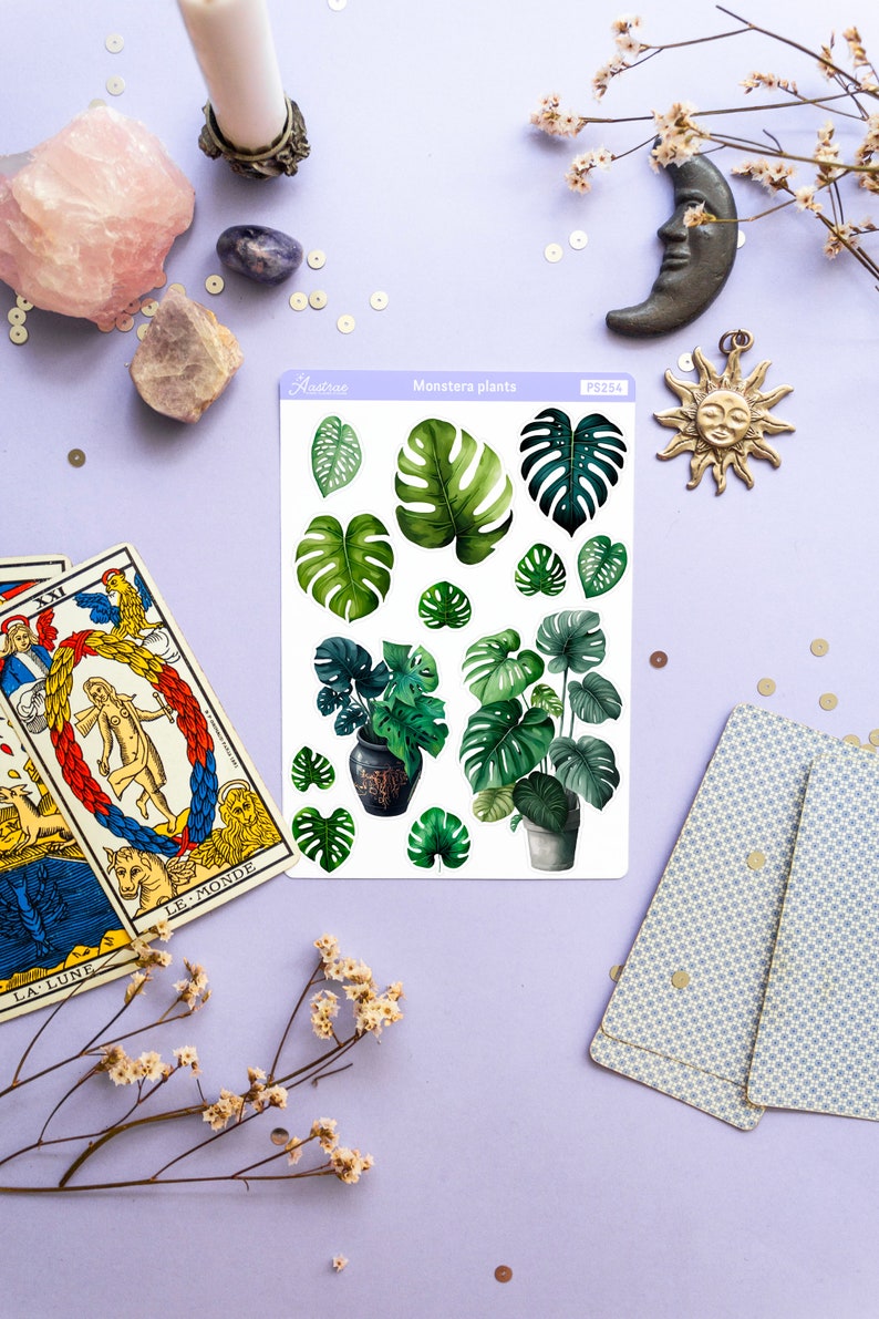 Puede incluir: Una hoja de pegatinas de plantas Monstera con varios dise&ntilde;os de hojas verdes y plantas en macetas. La hoja est&aacute; sobre una superficie de color lila claro, rodeada de cartas del tarot, cristales y objetos decorativos.