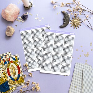 Op de afbeelding: Twee vellen met galaxy-kalender stickers met een rasterpatroon en een verloopachtergrond. De stickers bevinden zich op een paarse ondergrond met gouden glitter, een roze kristal, een witte kaars, een tarotkaart en een decoratieve maan en zon.