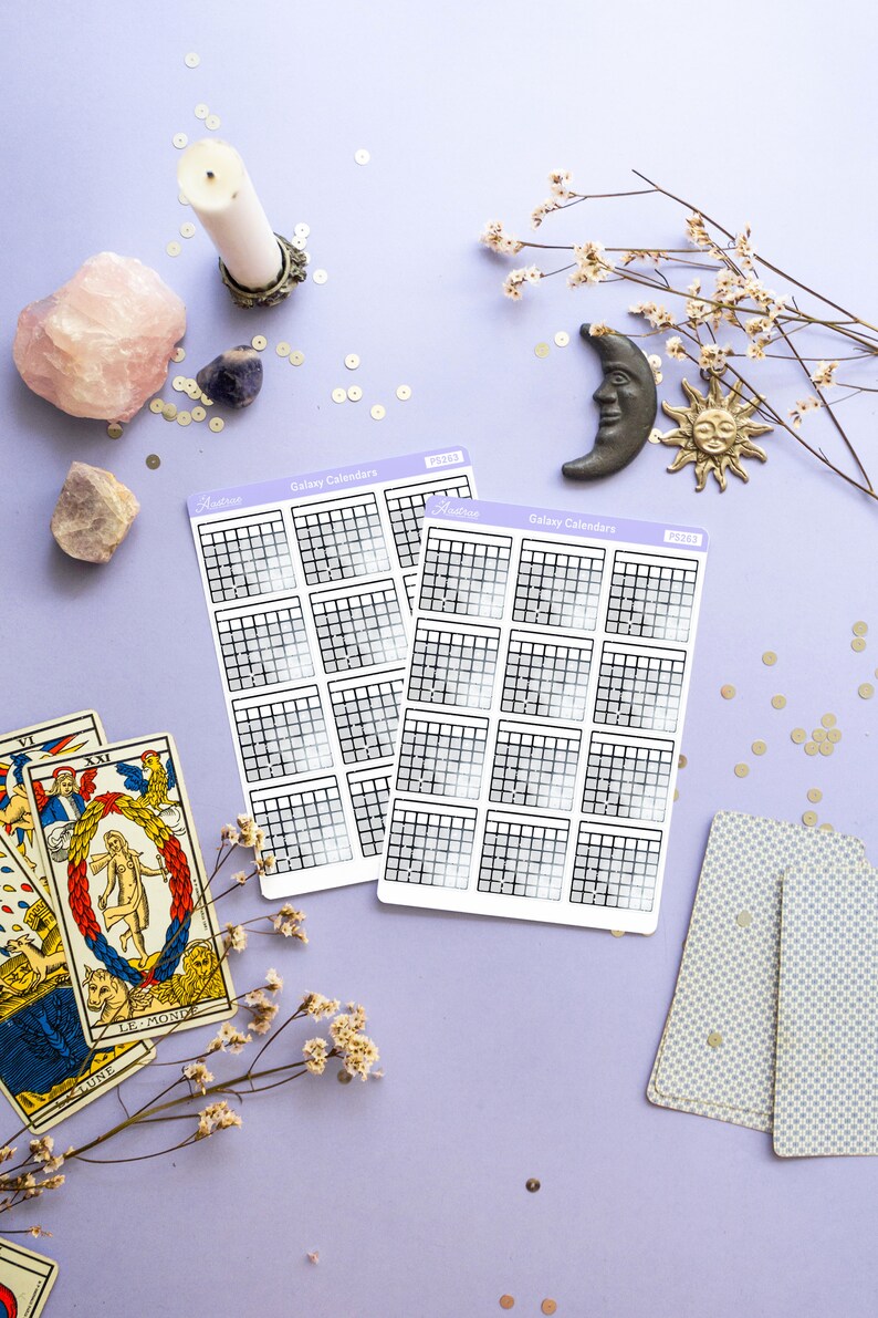 Op de afbeelding: Twee vellen met galaxy-kalender stickers met een rasterpatroon en een verloopachtergrond. De stickers bevinden zich op een paarse ondergrond met gouden glitter, een roze kristal, een witte kaars, een tarotkaart en een decoratieve maan en zon.