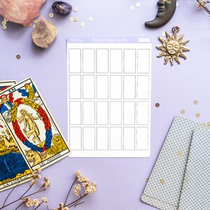 May include: A flat lay featuring tarot cards, blank tarot cards, crystals, and decorative elements on a lavender surface. The blank cards are rectangular with a grid of boxes. Other items include a candle, moon, and sun charms.