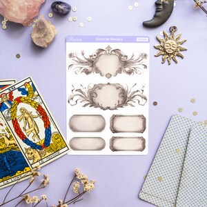 K&ouml;nnte beinhalten: Eine Blatt mit viktorianischen Banner-Aufklebern mit verzierten Rahmen und leeren Bannern. Die Aufkleber befinden sich auf einem violetten Hintergrund mit Tarotkarten, Kristallen und Dekorationsartikeln.