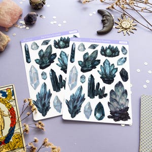 May include: Two sticker sheets with dark blue crystal illustrations, labeled "Dark Fairy Crystals", are displayed on a light purple surface. Decorative items include a candle, moon and sun charms, dried flowers, and a tarot card.