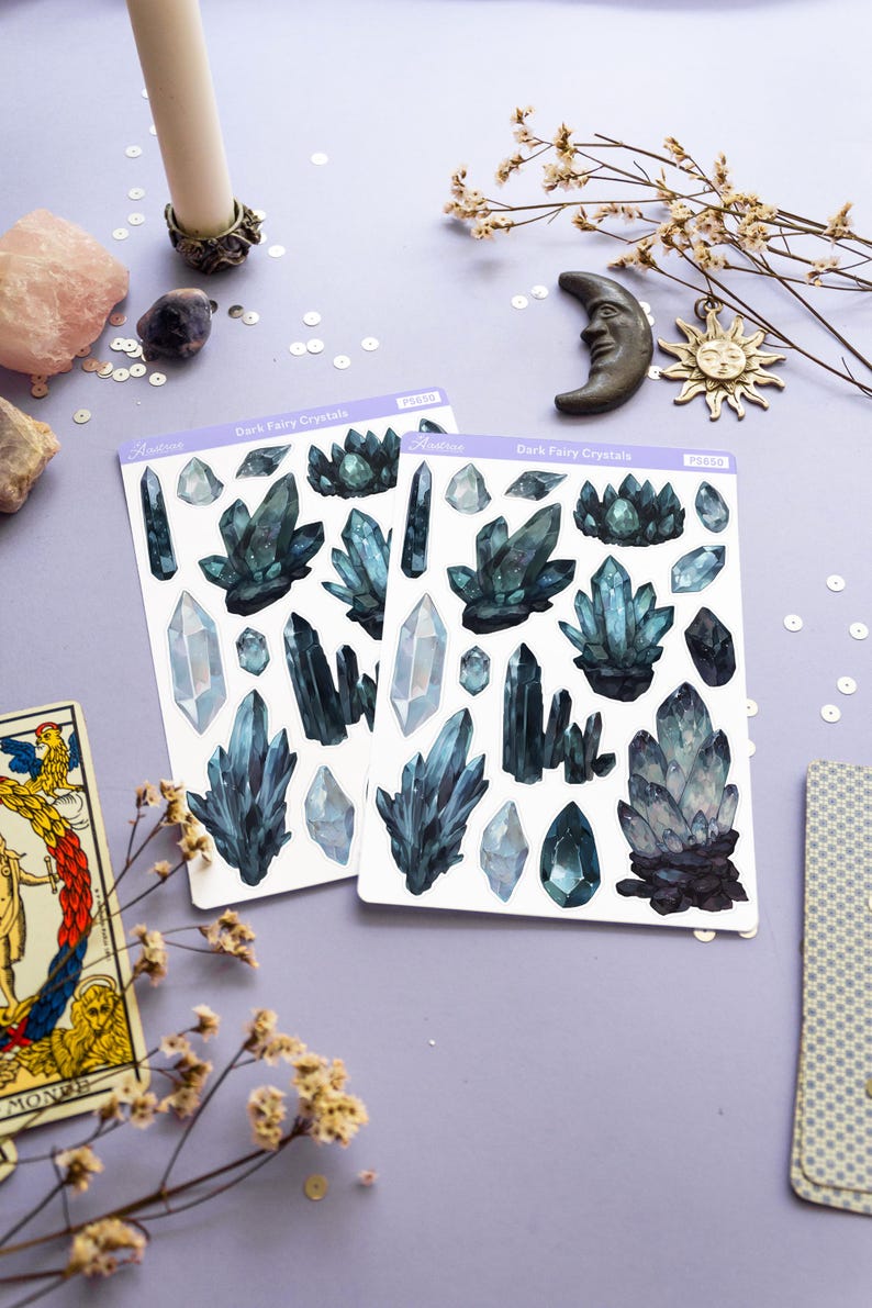 May include: Two sticker sheets with dark blue crystal illustrations, labeled "Dark Fairy Crystals", are displayed on a light purple surface. Decorative items include a candle, moon and sun charms, dried flowers, and a tarot card.