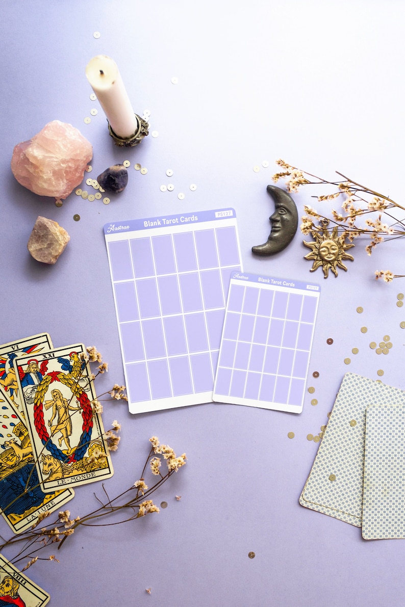 May include: A still-life arrangement featuring tarot cards, blank tarot card sheets, crystals, a candle, and celestial-themed decor on a lavender surface. The blank cards have a grid design. Dried flowers and gold sequins add detail.