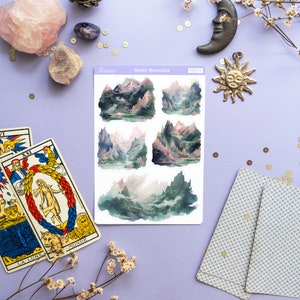 May include: A sheet of stickers with watercolor illustrations of misty mountains. The stickers are on a white background and have the text "Mystic Mountains" and "Astr&eacute;e" printed on them. The stickers are surrounded by tarot cards, a candle, crystals, and other decorative items.
