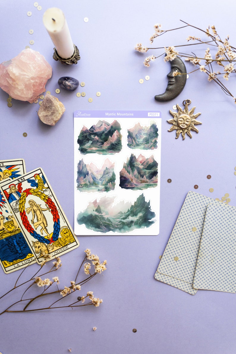 May include: A sheet of stickers with watercolor illustrations of misty mountains. The stickers are on a white background and have the text "Mystic Mountains" and "Astr&eacute;e" printed on them. The stickers are surrounded by tarot cards, a candle, crystals, and other decorative items.