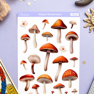 Peut inclure: Une feuille d'autocollants repr&eacute;sentant 15 illustrations de diff&eacute;rents champignons rouges et orange avec des points blancs. Les autocollants sont sur un fond blanc et la feuille est intitul&eacute;e "Champignons magiques" et "PS252".