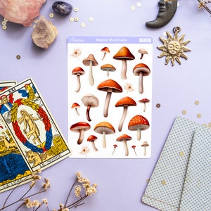 Peut inclure: Une feuille d'autocollants illustrant diff&eacute;rents types de champignons. Les autocollants sont sur fond blanc et ont une bordure rose clair. Le texte "Magical Mushrooms" est imprim&eacute; en haut de la feuille. La feuille est pos&eacute;e sur une surface violet clair avec divers objets d&eacute;coratifs, dont des cartes de tarot, des cristaux et un petit soleil en m&eacute;tal.