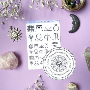 May include: A white card with black text and symbols depicting the eight pagan festivals. The card is on a purple background with white flowers, a pink crystal, a green crystal, a gray moon, and a silver sun charm.