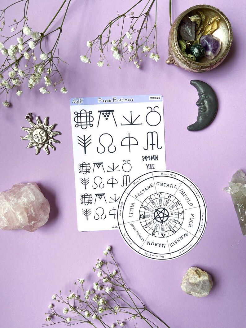 May include: A white card with black text and symbols depicting the eight pagan festivals. The card is on a purple background with white flowers, a pink crystal, a green crystal, a gray moon, and a silver sun charm.