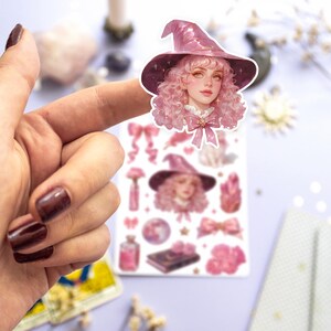 May include: A sheet of pink and white stickers featuring a witch with pink hair and a sparkly pink hat. The stickers also include other witchy elements like stars, moons, bottles, and flowers.