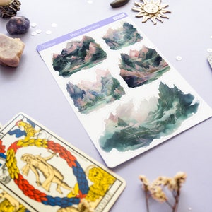 May include: A sheet of stickers featuring five watercolor illustrations of misty mountains. The stickers are on a white background and have a purple border. The text "Mystic Mountains" is printed on the sheet.