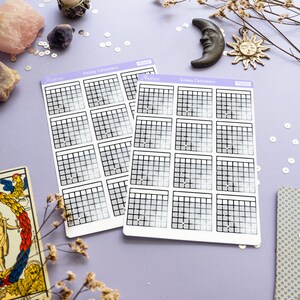 Op de afbeelding: Twee vellen met galaxy-kalender stickers met een holografische afwerking. Elk vel heeft 12 maandelijkse kalenders met een rasterindeling. De stickers bevinden zich op een paarse achtergrond met decoratieve items zoals een kaars, een maan, een zon en bloemen.