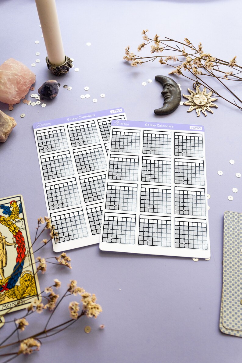 Op de afbeelding: Twee vellen met galaxy-kalender stickers met een holografische afwerking. Elk vel heeft 12 maandelijkse kalenders met een rasterindeling. De stickers bevinden zich op een paarse achtergrond met decoratieve items zoals een kaars, een maan, een zon en bloemen.