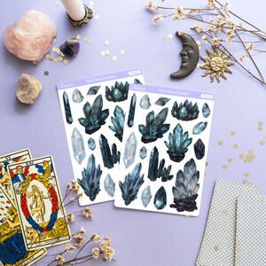 May include: Two sticker sheets with dark blue crystal designs, labeled "Dark Fairy Crystals", are displayed on a lavender surface. Other items include tarot cards, crystals, a candle, and decorative celestial objects.