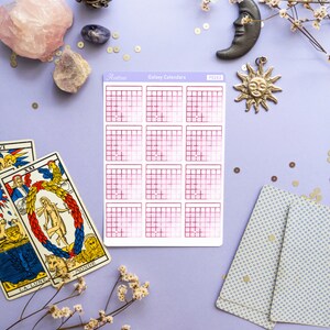 Op de afbeelding: Een vel roze en witte galaxy-kalender stickers met de tekst "Astro" en "Galaxy Calendars" gedrukt op de bovenkant. De stickers liggen op een paarse ondergrond met tarotkaarten, kristallen en decoratieve items.
