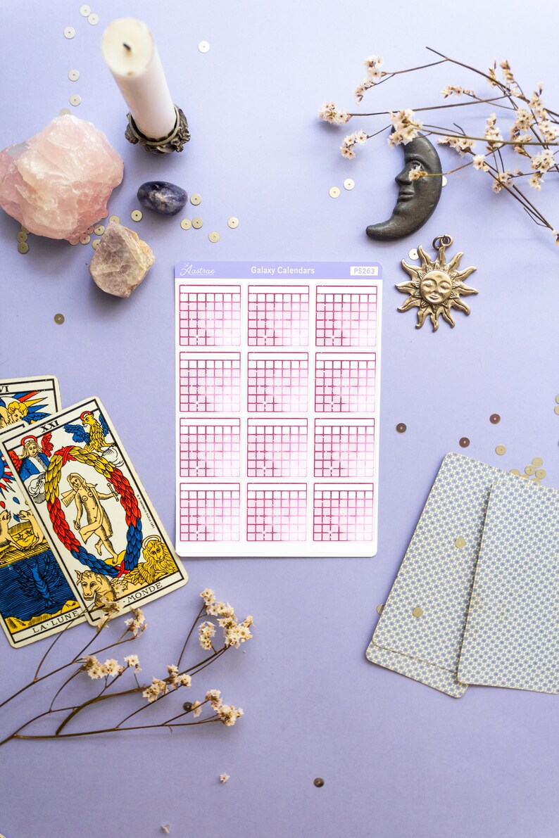 Op de afbeelding: Een vel roze en witte galaxy-kalender stickers met de tekst "Astro" en "Galaxy Calendars" gedrukt op de bovenkant. De stickers liggen op een paarse ondergrond met tarotkaarten, kristallen en decoratieve items.