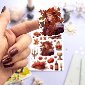 May include: A sheet of stickers featuring a woman with long red hair wearing a crown of autumn leaves. The stickers also include images of apples, candles, and a bottle of wine.