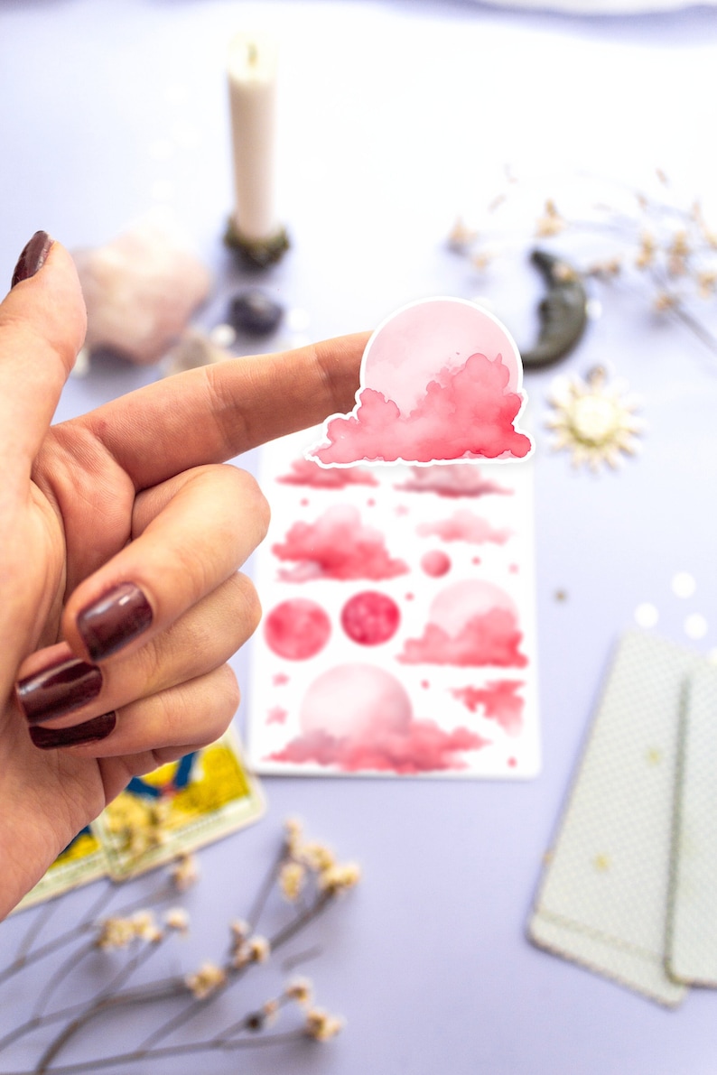 May include: A sheet of pink watercolor stickers featuring various moon and cloud designs. The stickers are perfect for adding a touch of whimsy to journals, planners, and other craft projects.