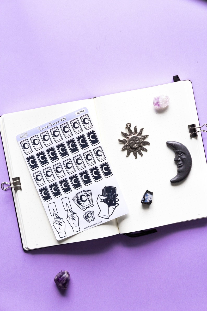 May include: A sheet of stickers featuring tarot cards with a crescent moon and stars design. The stickers are on a white background and are arranged in rows. There are also stickers of hands holding tarot cards. The stickers are on a notebook with a purple background.