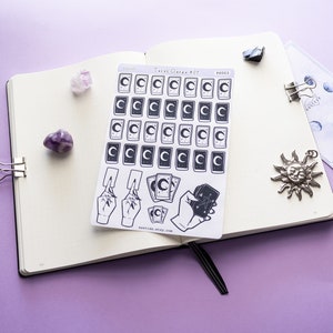 May include: A sheet of stickers featuring tarot card designs with a crescent moon and stars. The stickers are on a white background and are arranged in a grid pattern. There are also stickers of hands holding tarot cards. The stickers are on a purple surface with other decorative items.