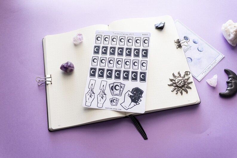 May include: A sheet of stickers featuring tarot card designs with a crescent moon and stars. The stickers are on a white background and are arranged in a grid pattern. There are also stickers of hands holding tarot cards. The stickers are on a purple surface with other decorative items.