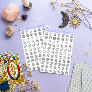 May include: Two sheets of stickers with witch icons. The stickers are white with black outlines and feature icons such as moons, stars, crystals, cauldrons, and books. The stickers are on a purple background with tarot cards, flowers, and crystals.